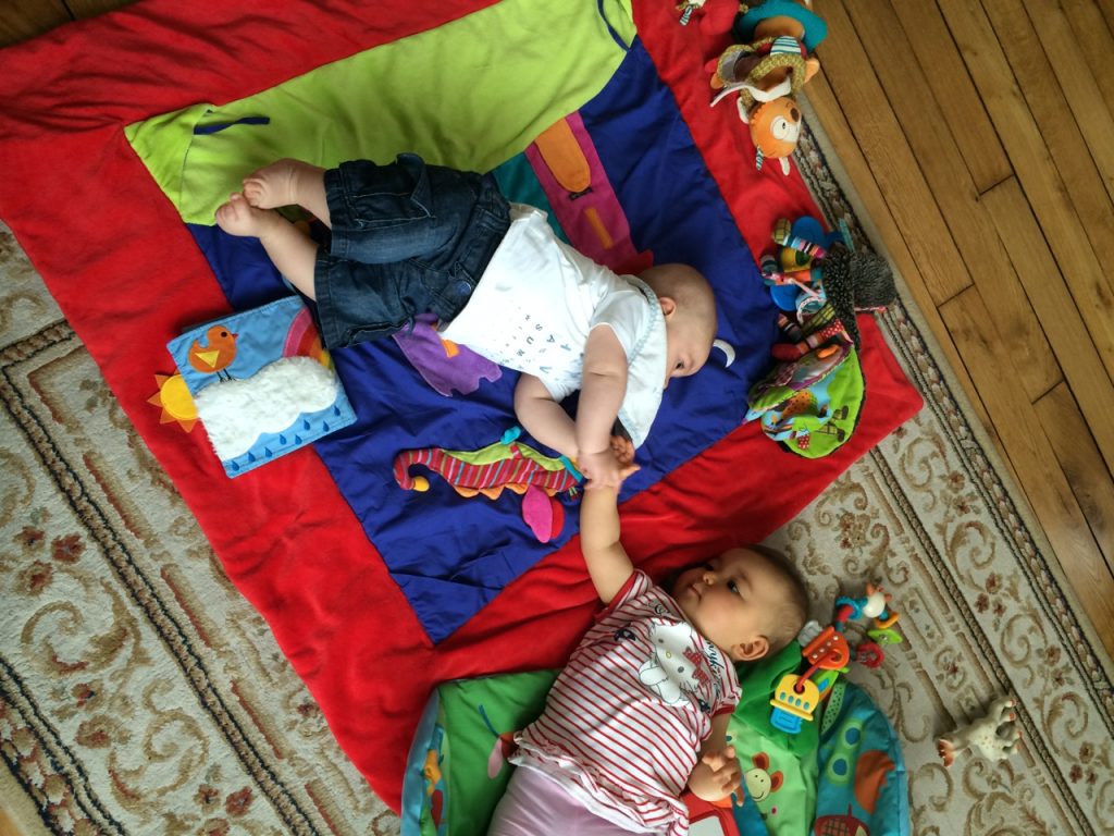 Deux bébés allongés sur des tapis de jeu qui se touchent les mains.