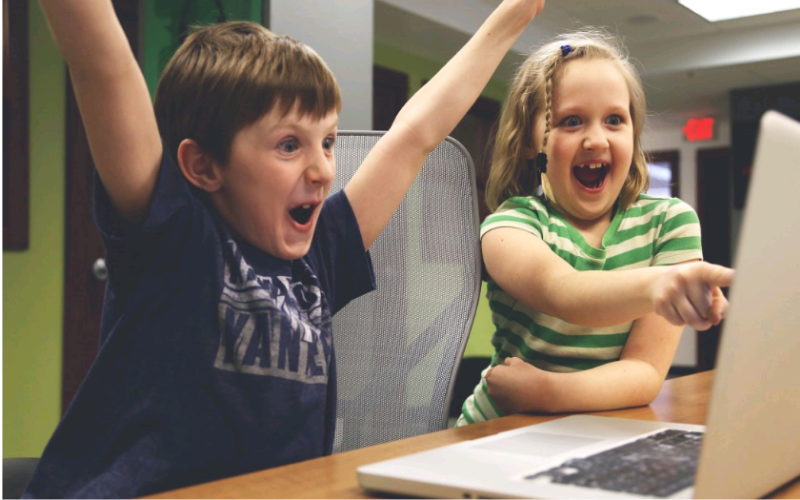 Deux enfants devant un ordinateur. L'un lève les bras, l'autre pointe l'écran en riant. 