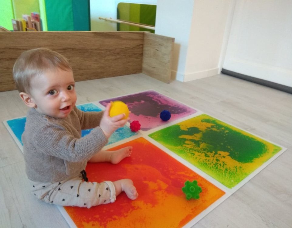 Jeune enfant assis sur des plaques sensori-motrices et qui tient une balle jaune dans ses mains.