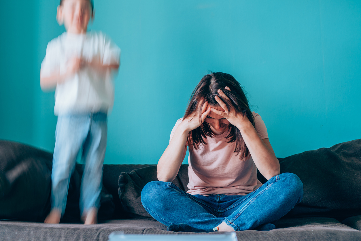 Femme qui se tient la tête sur un canapé pendant que son fils saute sur celui-ci en arrière plan.