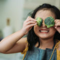 Une enfant tient deux fleurons de brocolis devant ses yeux en souriant.