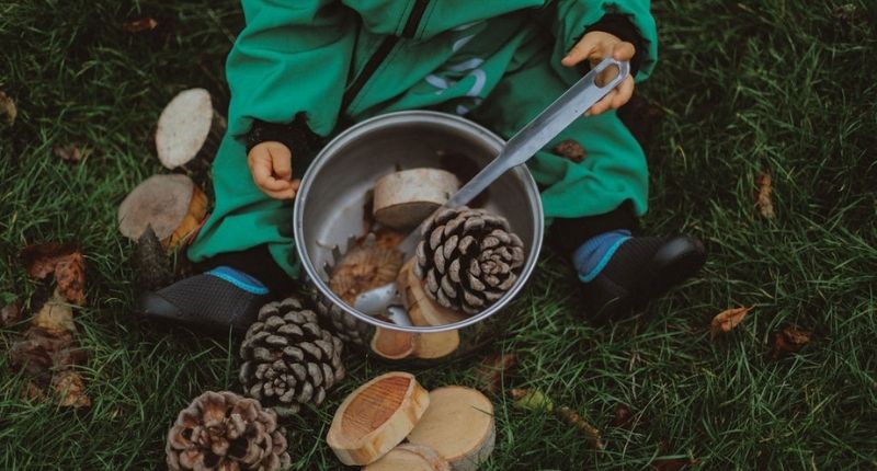 Enfant qui joue assis dans l'herbe avec des morceaux de bois, de pommes de pin dans une casserole.