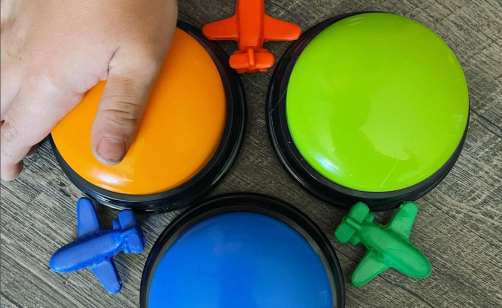 Une personne appuie sur un buzzer orange. Il y aussi un buzzer vert et un bleu.