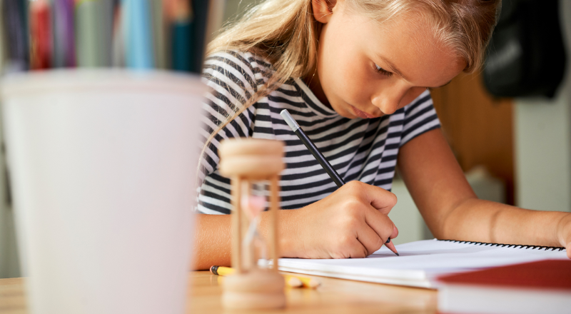 Un enfant fait ses devoirs. Un sablier en bois s'écoule devant elle.