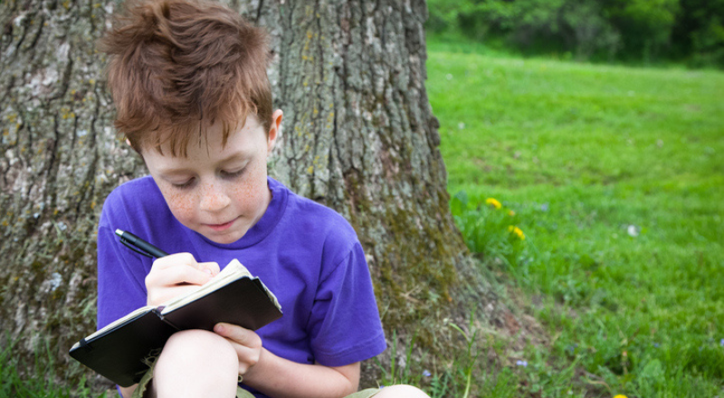 Un niño apoyado en un árbol escribe en un cuaderno negro