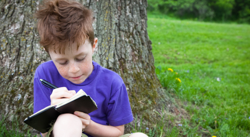 Un enfant adossé contre un arbre écrit dans un carnet noir