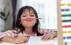 une petite fille assise à un bureau qui joue avec son crayon en le faisant tenir en équilibre à l'horizontal au-dessus de sa bouche.