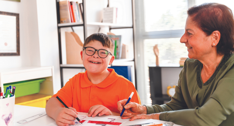 garçon trisomique qui apprend une leçon avec une dame. Les deux sourissent.