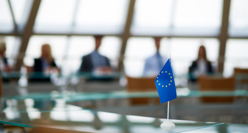 petit drapeau européen posé sur une table en verre où l'on aperçoit des personnes en flou derrière.