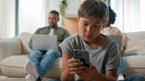 Enfant face à un smartphone, ses parents en arrière plan. Il semble préoccupé