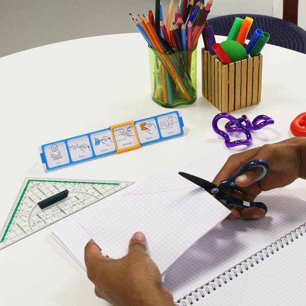 Mains d'enfant découpant un papier durant une activité créative avec un séquencier de routine en arrière-plan