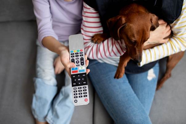 Deux personnes et un chien sur un canapé, l'une d'entre elles tient une télécommande